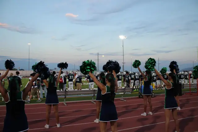 The squad cheering with poms raised along the track at the August 2026 home opener